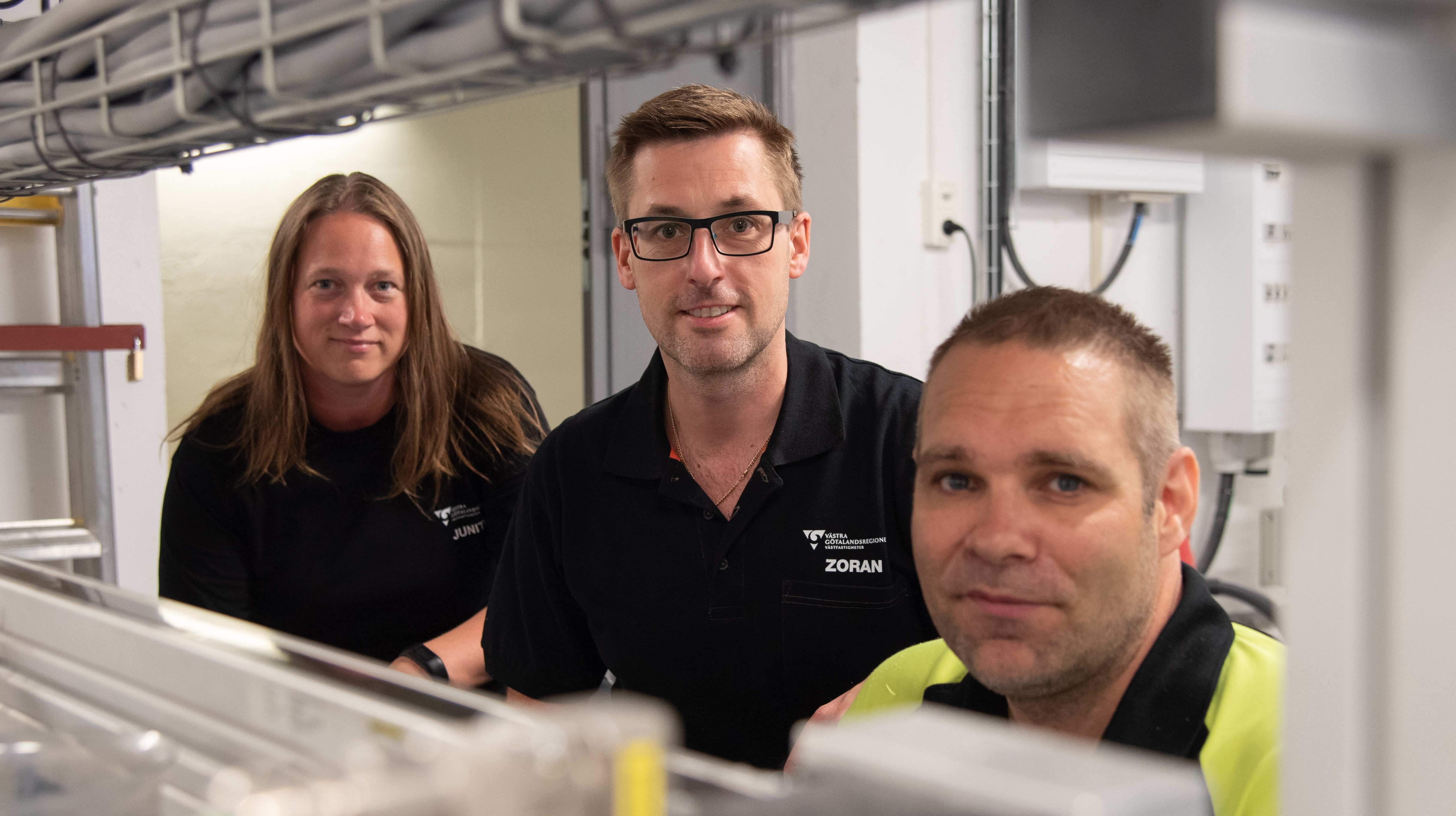 Tre människor som står bland rör och eluttag tittar in i kameran